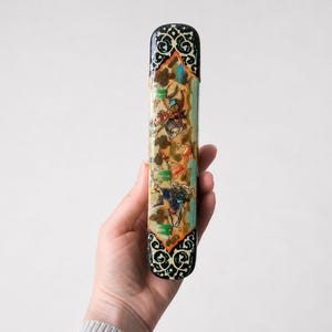 Hand holding a handcrafted Persian miniature pen holder featuring intricate painted artwork and ornate marquetry, photographed against a clean light grey studio background.