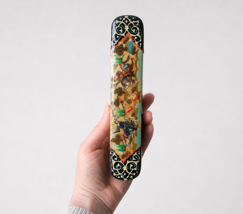 Hand holding a handcrafted Persian miniature pen holder featuring intricate painted artwork and ornate marquetry, photographed against a clean light grey studio background.