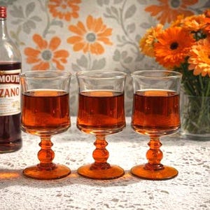 Puede incluir: Tres copas de tallo llenas de un líquido marrón rojizo, probablemente un cóctel, sobre un mantel de encaje. Una botella de vermut Cinzano a la izquierda. Un jarrón de flores naranjas en el fondo.