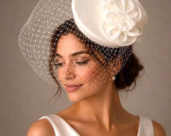 May include: A white fascinator hat featuring a circular base, a layered satin flower accent, and a delicate white birdcage veil with a diamond-shaped mesh pattern that covers the upper portion of the face.