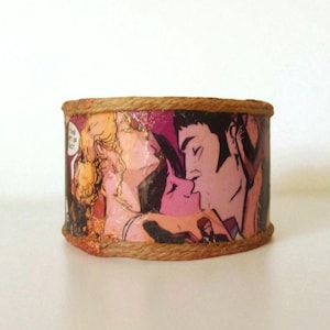 Op de afbeelding: Een cilindrische decoratieve container met een stripboekillustratie van een kussend stel. De rand is omwikkeld met een natuurlijk gekleurd touw. De stripkunst heeft roze, zwarte en gele tinten.
