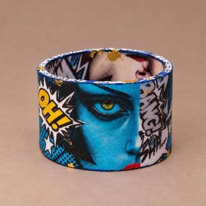 May include: A cylindrical cuff bracelet featuring a vibrant pop art graphic print. The design displays a close-up of a face with blue skin, a yellow eye, and red lips, accented by comic book speech bubbles containing the text OH! and BANG!.