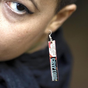May include: A pair of earrings with a silver hook and a rectangular charm. The charm is made of a comic book page with the word "CRYPTOID" printed in red and blue letters. The text on the charm reads "HELP!  YOU CAN MOVE!  IT'S A HELP!  YOU CAN MOVE!  IT'S A"