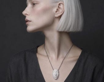 May include: A pendant necklace featuring a small, white, sculpted human face charm suspended from a thin, dark metallic chain. The charm is oval-shaped with detailed facial features including eyes, a nose, and lips, resting against a dark gray garment.