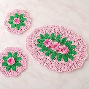 May include: Three-piece crochet set in pink, featuring a floral design. The set includes two coasters and an oval placemat. Each piece has a pink rose in the center, surrounded by green leaves, all on a pink background.
