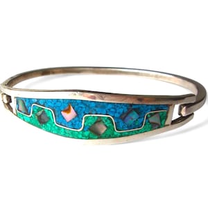 May include: Silver-tone hinged bangle bracelet with a turquoise and green inlay design. The inlay features a wave pattern with small turquoise stones and square-shaped accents in various colors, including pink and iridescent. A stylish piece of jewelry.