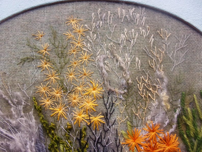 A detailed macro close-up of a hand-embroidered floral meadow study showcasing dense orange and yellow wool yarn sunburst blooms, fine white and green branching stitches, and tightly clustered French knots on a textured linen background, framed by a light blue oval mount.