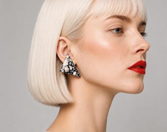 May include: A close-up of a woman wearing a white top and a triangular earring. The earring is black, white, and gray with a speckled pattern. The woman has short, blonde hair and red lipstick.