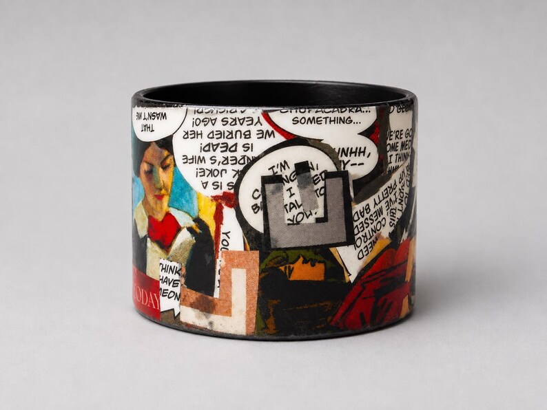 Handmade Pop Art comic bangle featuring a bold collage of illustrated characters, speech bubbles, an eye motif and red graphic details. Visible comic text includes “WE BURIED HER,” “IS DEAD,” and “THAT WASN’T ME.”