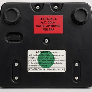 The black plastic base on the underside of a vintage telephone showing a red label and a green approved sticker with text.