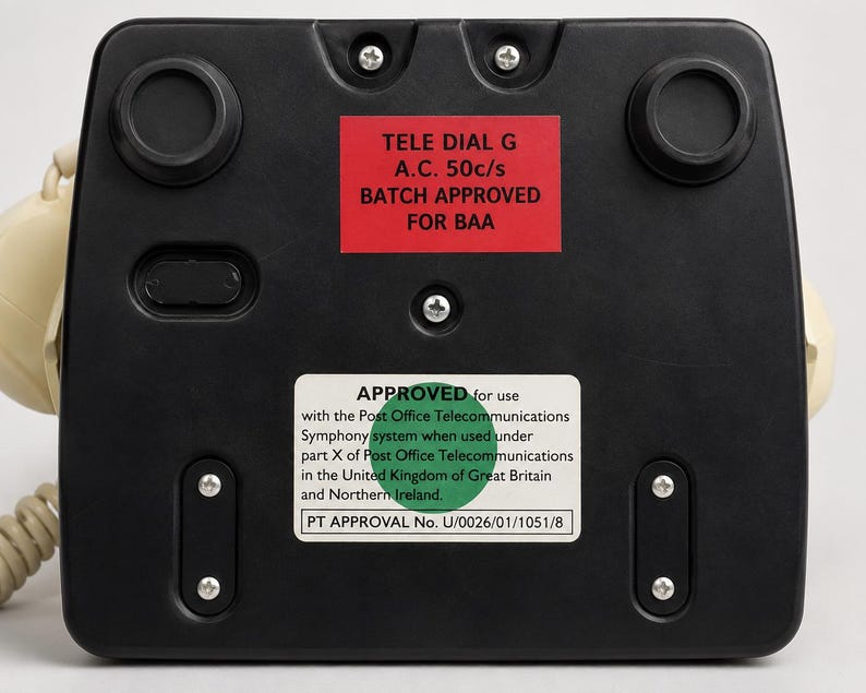 The black plastic base on the underside of a vintage telephone showing a red label and a green approved sticker with text.