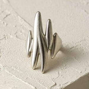 May include: A silver ring with an abstract design. The ring features a series of vertical, elongated shapes rising from the band, creating a unique aesthetic. The metal has a smooth, polished finish. The ring is a piece of jewellery.