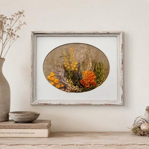A hand-embroidered oval floral meadow study with yellow and orange wool yarn sunburst blooms on a natural linen weave ground, presented inside a distressed light blue wooden frame resting on a rustic wooden console table against a neutral beige wall, flanked by vintage books, a ceramic vase with dried stems, and a small bird figurine next to a mossy nest with a speckled egg.