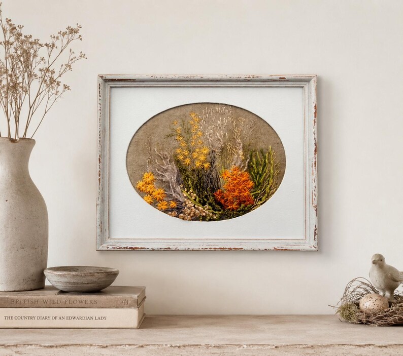 A hand-embroidered oval floral meadow study with yellow and orange wool yarn sunburst blooms on a natural linen weave ground, presented inside a distressed light blue wooden frame resting on a rustic wooden console table against a neutral beige wall, flanked by vintage books, a ceramic vase with dried stems, and a small bird figurine next to a mossy nest with a speckled egg.