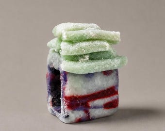 May include: A small, rectangular, hollow object with a textured, frosted surface. The base features a marbled pattern of white, deep red, and dark blue. Resting on top are several stacked, pale green, rectangular pieces with a powdery finish.