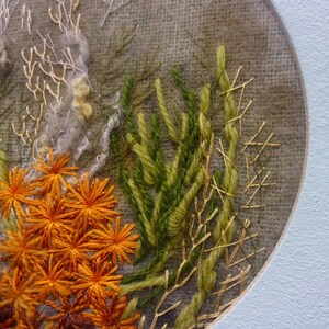 A detailed macro close-up of a hand-embroidered floral meadow study showcasing dense orange and yellow wool yarn sunburst blooms, fine white and green branching stitches, and tightly clustered French knots on a textured linen background, framed by a light blue oval mount.