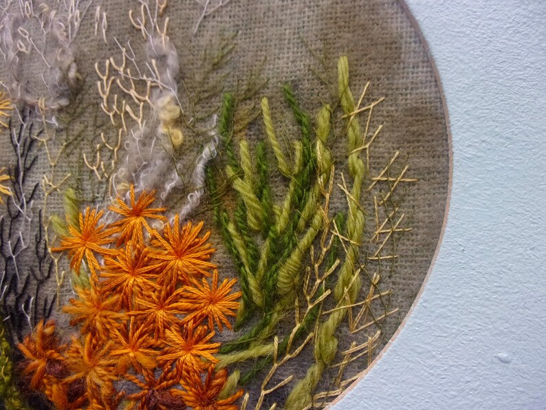 A detailed macro close-up of a hand-embroidered floral meadow study showcasing dense orange and yellow wool yarn sunburst blooms, fine white and green branching stitches, and tightly clustered French knots on a textured linen background, framed by a light blue oval mount.