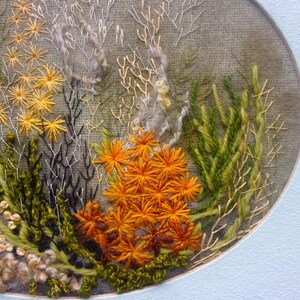 A detailed macro close-up of a hand-embroidered floral meadow study showcasing dense orange and yellow wool yarn sunburst blooms, fine white and green branching stitches, and tightly clustered French knots on a textured linen background, framed by a light blue oval mount.