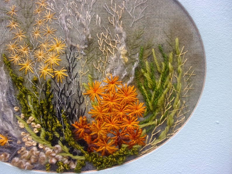 A detailed macro close-up of a hand-embroidered floral meadow study showcasing dense orange and yellow wool yarn sunburst blooms, fine white and green branching stitches, and tightly clustered French knots on a textured linen background, framed by a light blue oval mount.