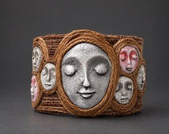 Pode incluir: Uma pulseira larga com uma base de juta castanha texturizada, adornada com vários rostos esculpidos. Cada rosto é emoldurado por uma corda castanha, com alguns acabamentos em cinzento pedra e outros com detalhes em tons de rosa e vermelho.