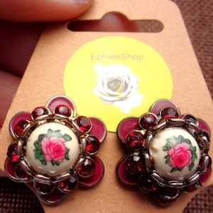 May include: A pair of red and gold earrings with a white centre featuring a floral design. The earrings are shaped like a flower with red rhinestones around the edge. The centre of the earrings has a white background with a pink rose and green leaves.