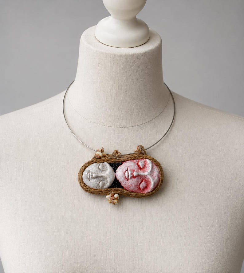 Two clay faces hand-shaped into an oval pendant bound by brown twine on a silver wire choker.