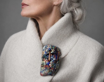 May include: A textured, rectangular brooch featuring a multi-colored mosaic pattern of blue, green, and red flecks, accented by thin, vertical copper-toned metal wires, pinned to the lapel of a thick, cream-colored wool coat with a high collar.