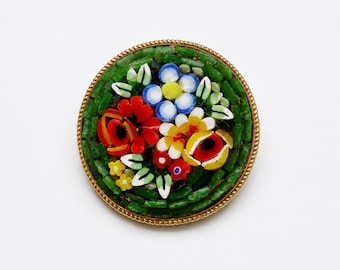 May include: A round brooch featuring a micro-mosaic floral design set in a gold-toned twisted metal frame. The center displays small glass tiles in red, yellow, blue, and white forming flowers, surrounded by a dense background of green glass tiles.