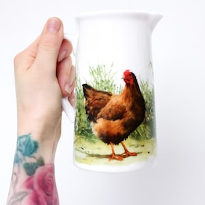 May include: White ceramic pitcher with a handle, featuring a detailed illustration of a brown hen with a red comb and wattle. The hen stands in a grassy field. The pitcher is a kitchenware item.