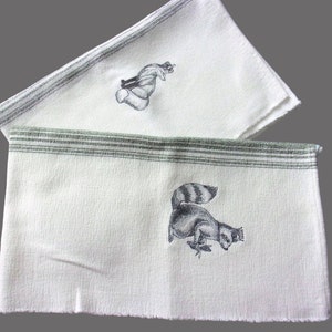 May include: Two white kitchen towels with green stripes. One towel features an embroidered image of a raccoon, the other features an embroidered image of a squirrel.