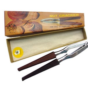 Può includere: Un schiaccianoci vintage con manico in legno e meccanismo di rottura in metallo argentato. Lo schiaccianoci è nella sua scatola originale, che è marrone con un'immagine di noci su di essa. La scatola dice "NUT CRACKER" sul davanti.