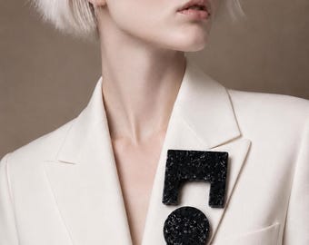 May include: A black brooch with a speckled texture, featuring a square-shaped top piece with a cutout and a circular bottom piece, pinned to the lapel of a crisp white blazer. The accessory has a glossy finish and a distinct geometric design.