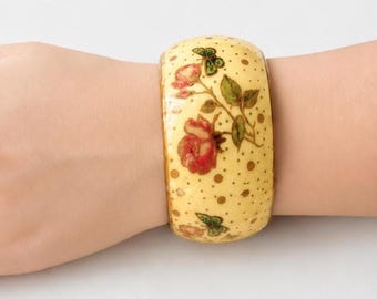 Może przedstawiać: Szeroka bransoletka typu bangle o kremowej podstawie, ozdobiona wzorem w brązowe kropki, czerwone kwiaty róż, zielone liście i małe zielone motyle. Bransoletka ma gładką, zaokrągloną powierzchnię oraz grubą i solidną konstrukcję.