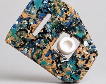 May include: Abstract geometric sculpture with a rectangular shape and a central opening. The sculpture features a speckled pattern of blue, orange, black, and white. A small, white, circular object is set within the central opening.