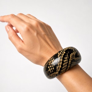 Può includere: Un bracciale rigido largo e curvo con finitura lucida e motivo a pelle di serpente nero e beige. Il bracciale presenta una superficie esterna liscia e arrotondata con una struttura spessa e solida che avvolge il polso.