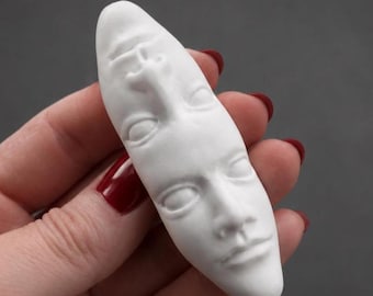 May include: A white, elongated, sculptural object featuring two human faces oriented in opposite directions. The faces are sculpted in relief with visible eyes, noses, and mouths, sharing a single continuous form with a smooth, matte finish.
