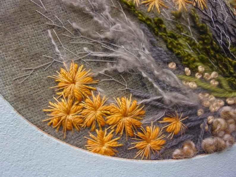 A detailed macro close-up of a hand-embroidered floral meadow study showcasing dense orange and yellow wool yarn sunburst blooms, fine white and green branching stitches, and tightly clustered French knots on a textured linen background, framed by a light blue oval mount.