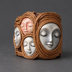 May include: A cylindrical vessel wrapped in thick brown rope, featuring four sculpted faces with closed eyes. Three faces are grey with a stone-like texture, while one smaller face is a reddish-pink hue. The faces are framed by the coiled rope.