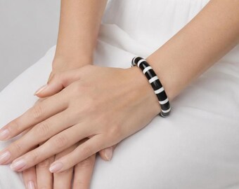 May include: A circular bangle bracelet featuring a repeating pattern of alternating thick black and thin white vertical stripes. The bracelet has a smooth, glossy finish and a rounded profile, encircling the wrist with a consistent color design.