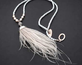 May include: A long necklace featuring light blue beads, a cluster of white pearls, and a grey stone bead. A large silver-toned metal cap secures a long, wispy grey feather tassel. The necklace includes a silver-toned oval toggle clasp and a small oval charm.