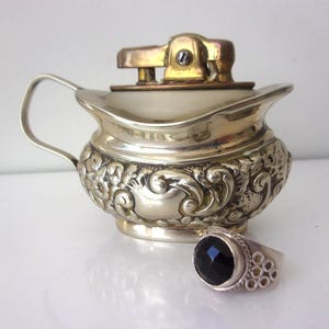 May include: A vintage silver-toned lighter with a decorative floral pattern and a gold-colored top. A silver ring with a large black gemstone sits next to the lighter. The lighter has a handle.
