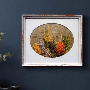 A hand-embroidered oval floral meadow study with yellow and orange wool yarn sunburst blooms on a natural linen weave ground, presented inside a distressed light blue wooden frame resting against a dark navy blue wall.