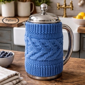 May include: A stainless steel French press coffee maker with a blue knitted cozy featuring a cable knit design. The cozy covers the glass carafe, providing insulation. The coffee maker sits on a wooden surface.