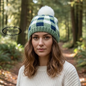 May include: A knitted hat in a green, white, and navy blue plaid pattern, topped with a white pom-pom. The person in the image is wearing a cream-coloured sweater. The word "Pattern" is in a black oval on the left side of the image.