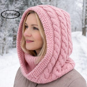 May include: A pink knitted hood and cowl set. The hood has a cable knit design on the front and a ribbed texture on the sides. The word "Pattern" is in a white oval in the upper left corner. The cowl is around the neck.