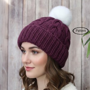 May include: A knitted plum-colored beanie with a cable knit design and a white pom-pom. The hat is worn by a person. The word "Pattern" is in a small oval on the right side of the image. The background is a light brown wooden wall.