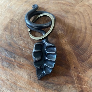 May include: A dark grey, hand-forged metal keyring featuring a textured leaf-shaped pendant attached to a coiled, spiral-shaped metal top. A circular brass-toned metal split ring is threaded through the spiral section of the keyring.