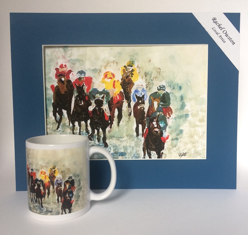 May include: A white ceramic mug with a colorful painting of horses and riders on a foggy racetrack. The mug is next to a framed painting of the same scene, with the artist's name "Rachel Ouston, Local Artist" in the top right corner.