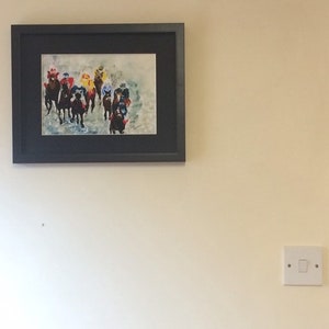 May include: A framed watercolor painting of a group of horses racing. The horses are in a line, with their heads down and their legs moving. The riders are wearing colorful jockey silks. The painting is set against a light blue background.