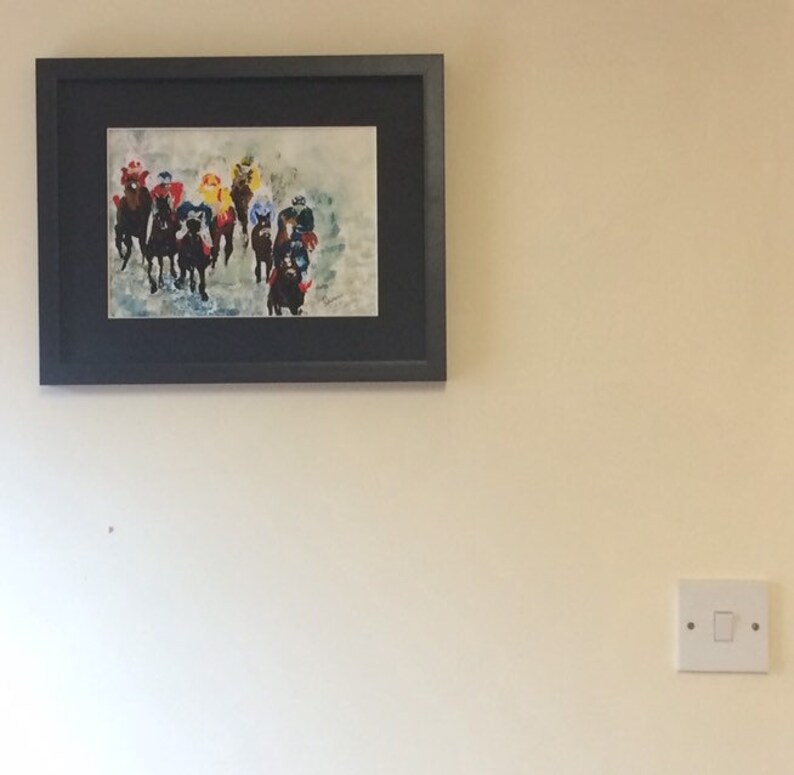 May include: A framed watercolor painting of a group of horses racing. The horses are in a line, with their heads down and their legs moving. The riders are wearing colorful jockey silks. The painting is set against a light blue background.